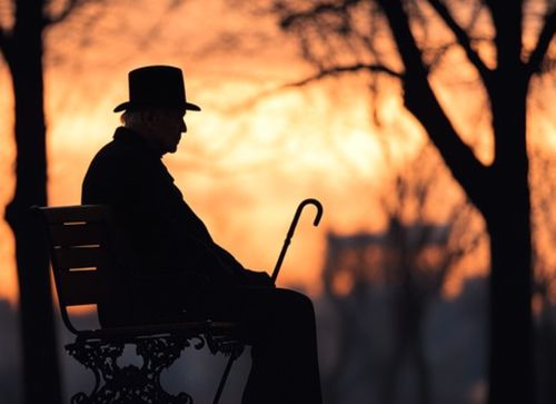 Elderly adult with a cane sitting on a bench at sunset